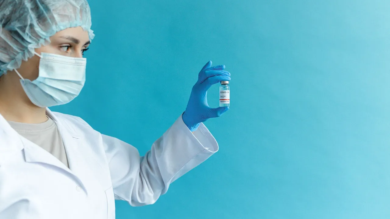 Female doctor in protective gear holding a vaccine vial against a blue background.