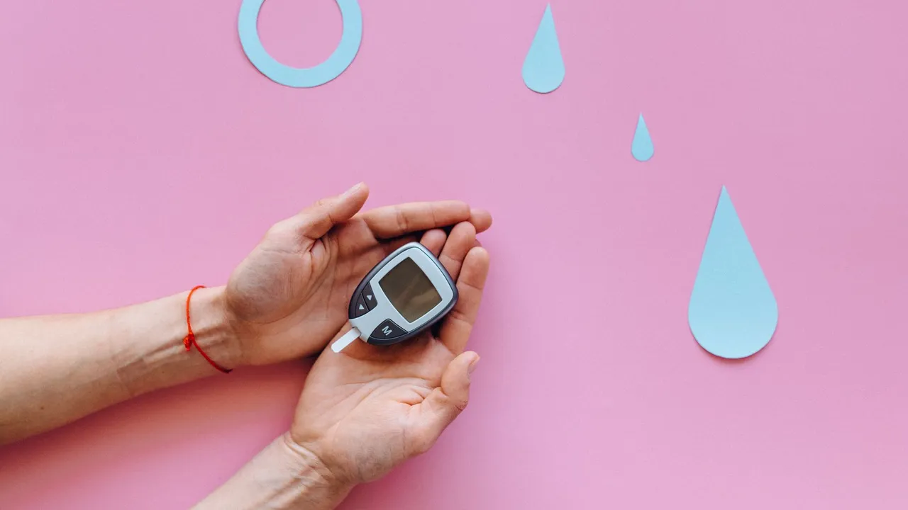 Hands holding a glucometer on a pink surface with blue diabetes symbols.