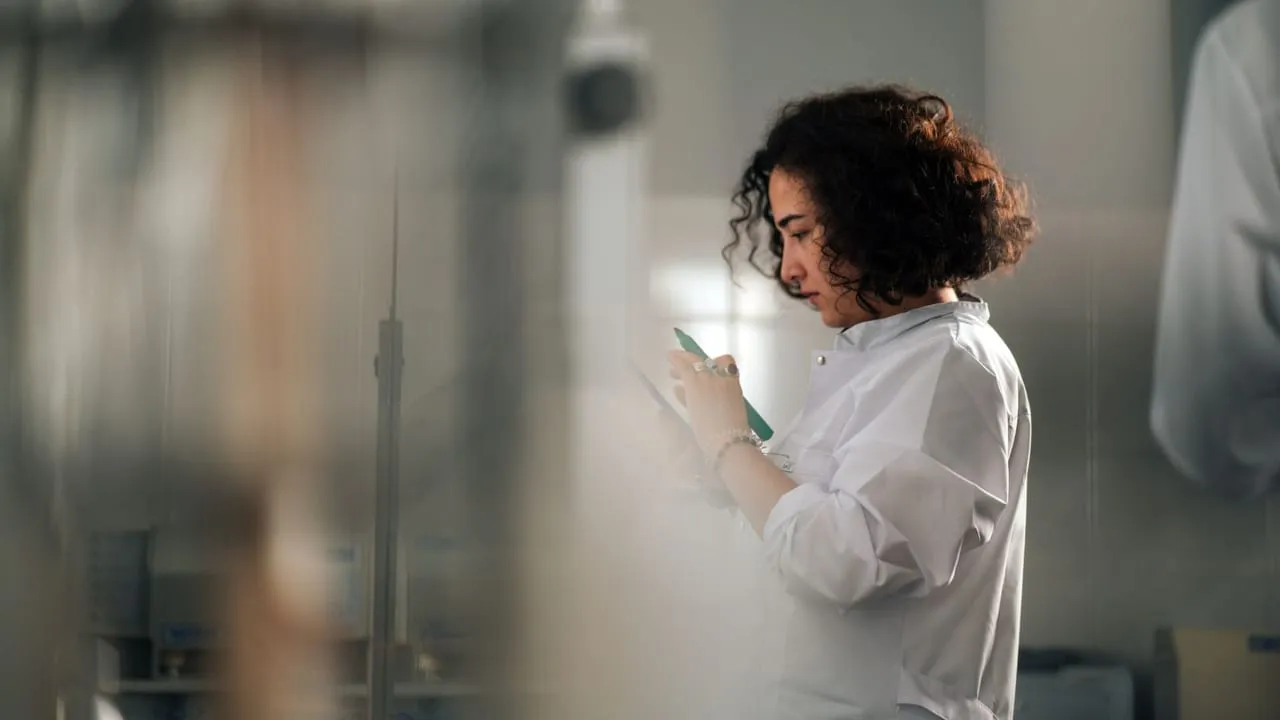 A focused female scientist with curly hair writes notes in a laboratory setting.
