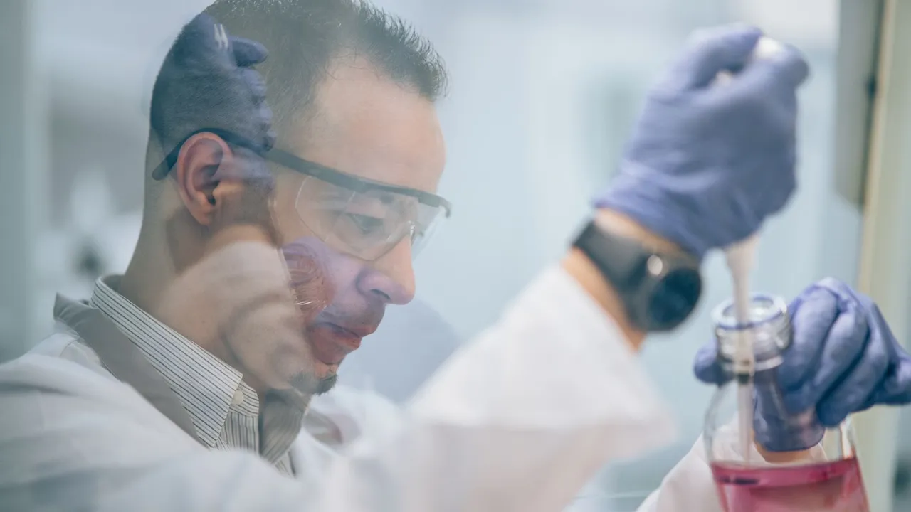 Scientist in lab coat and gloves transferring pink liquid using pipette.