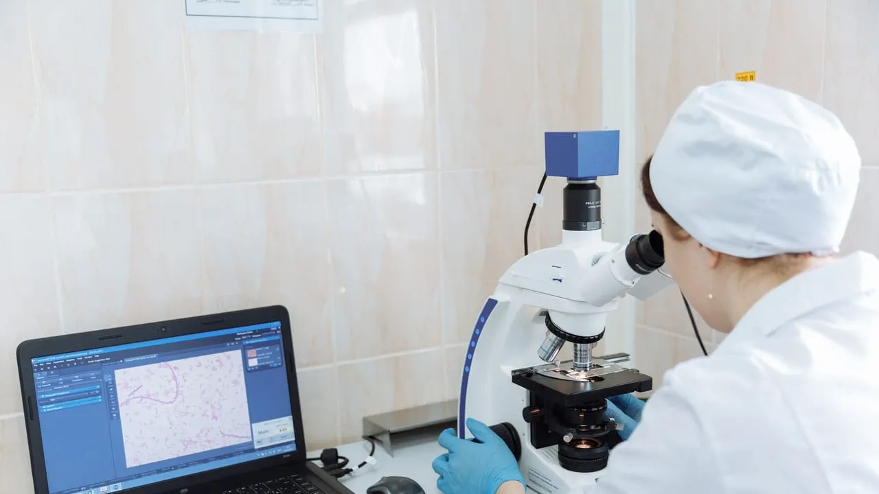Scientist in lab coat using microscope and laptop in a laboratory setting.