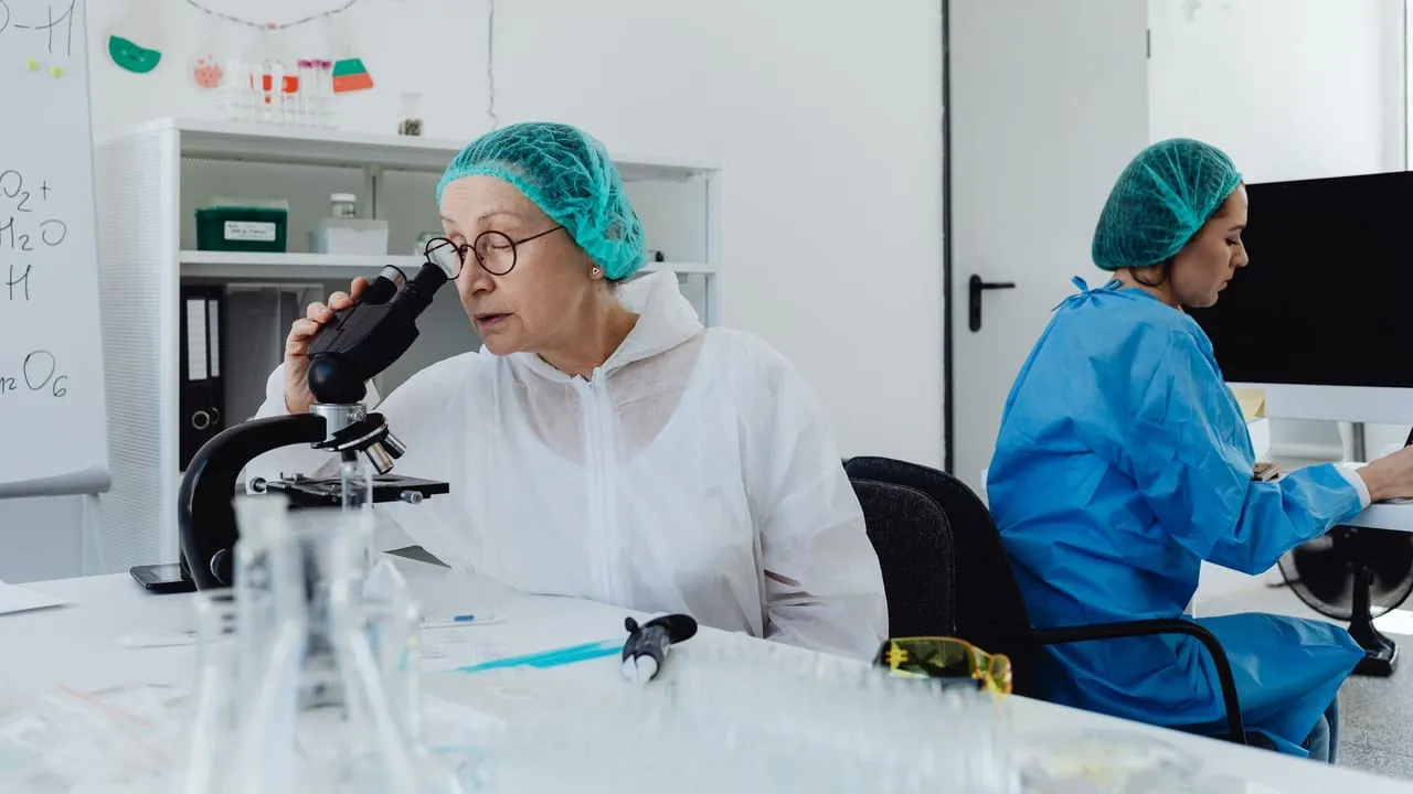 Scientists in coveralls and hair nets conducting research with microscopes and computers in a lab.