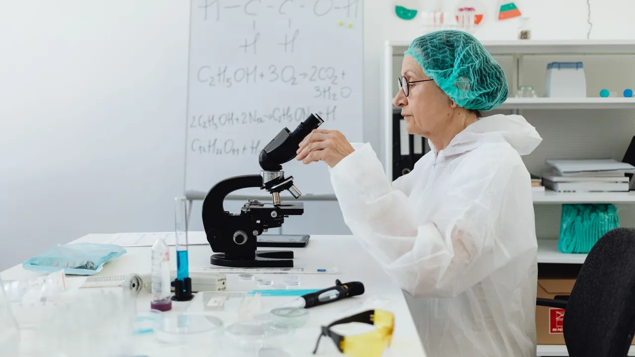 Senior scientist using a microscope in a laboratory setting wearing protective gear.