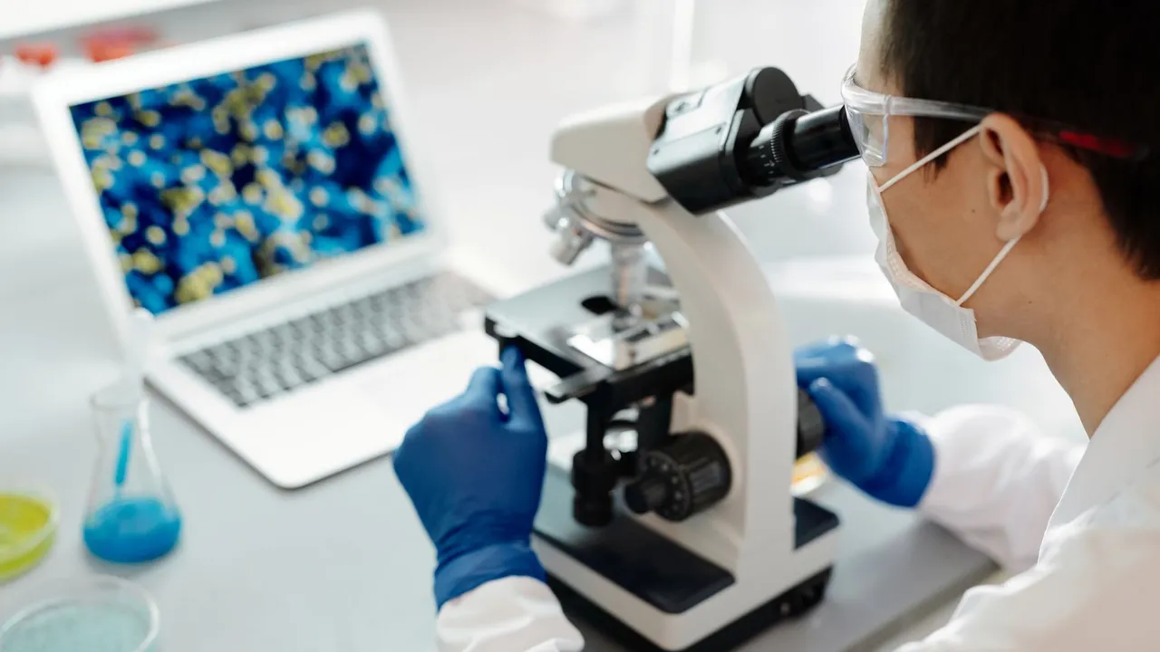 Scientist examining samples under a microscope in a lab with laptop display.