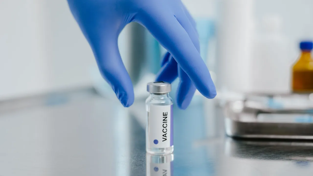 Close-up of a vaccine vial on a metal surface with a gloved hand in a laboratory setting.
