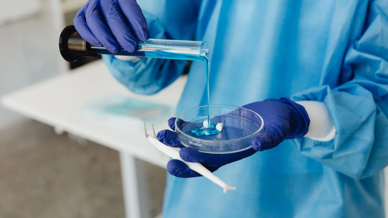 Scientist in lab gown pouring blue liquid from test tube into petri dish wearing gloves.