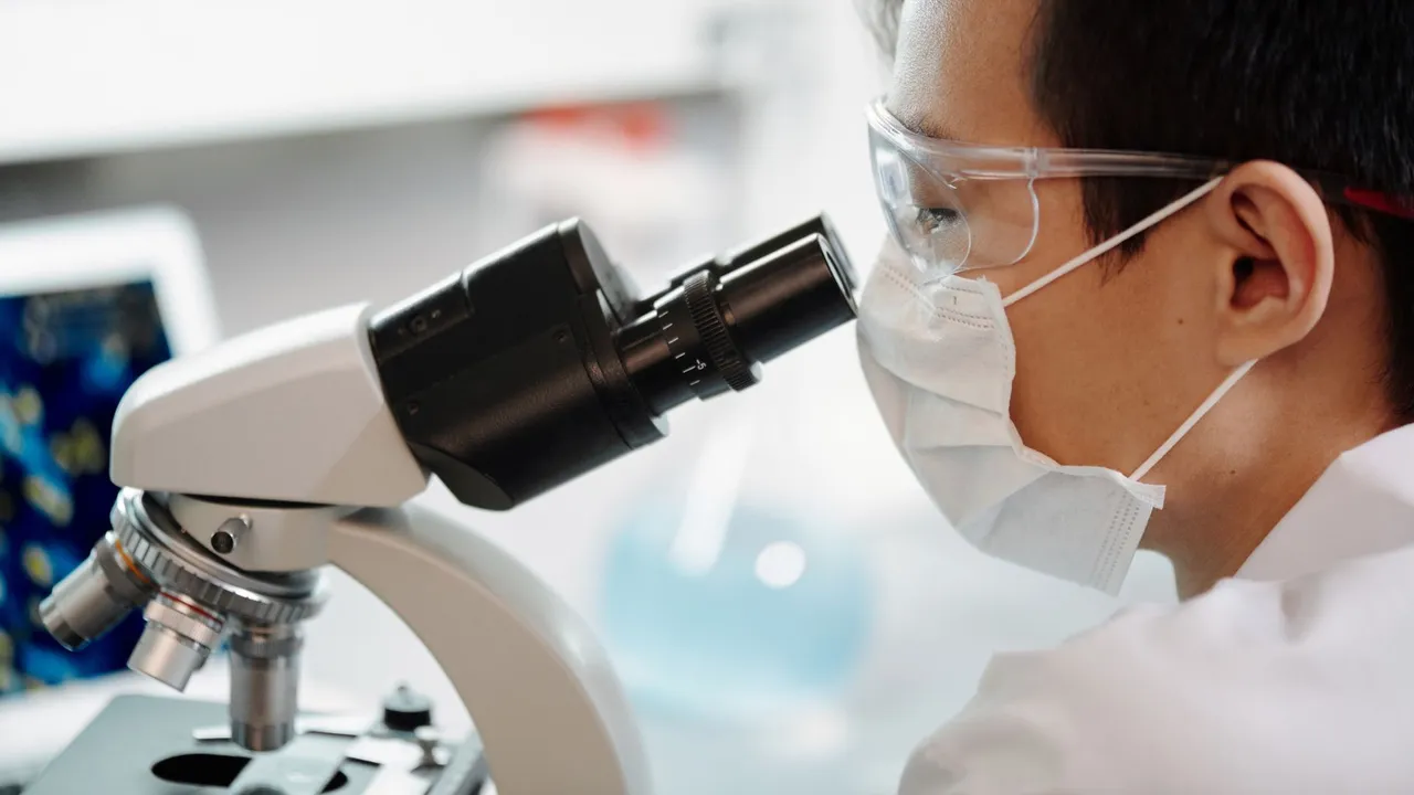 Scientist wearing face mask and goggles examining samples through a microscope in a lab.