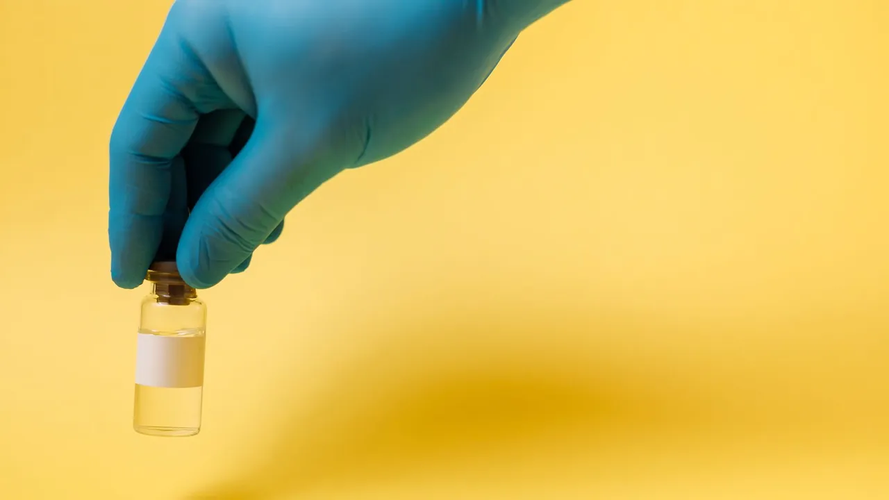 Close-up of a blue gloved hand holding a vaccine vial on a yellow background.