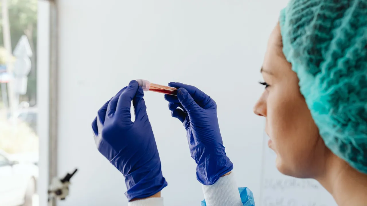 A focused lab technician holds a blood sample in a test tube, wearing gloves and a hair net.