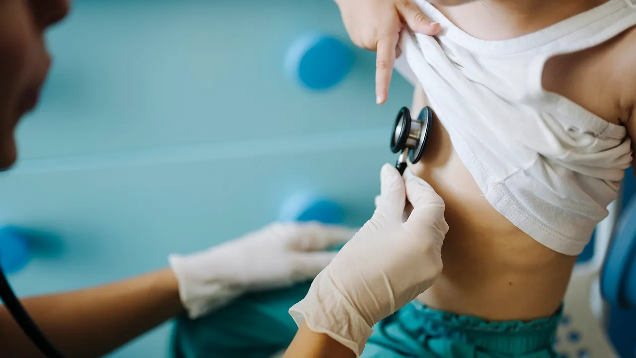 A doctor checks a child's heartbeat in a clinic setting. The child sits on a chair with a doctor's hand touching the stethoscope. The environment is bright and