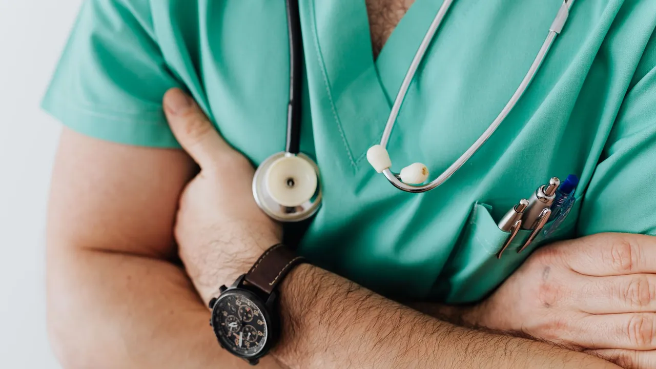 Unrecognizable man wearing medical uniform with stethoscope and pens standing in hospital with crossed arms during work in day time