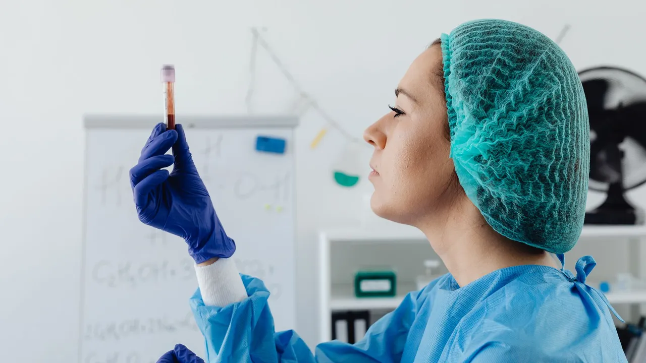 A female scientist in protective gear inspects a blood sample vial in a lab setting.