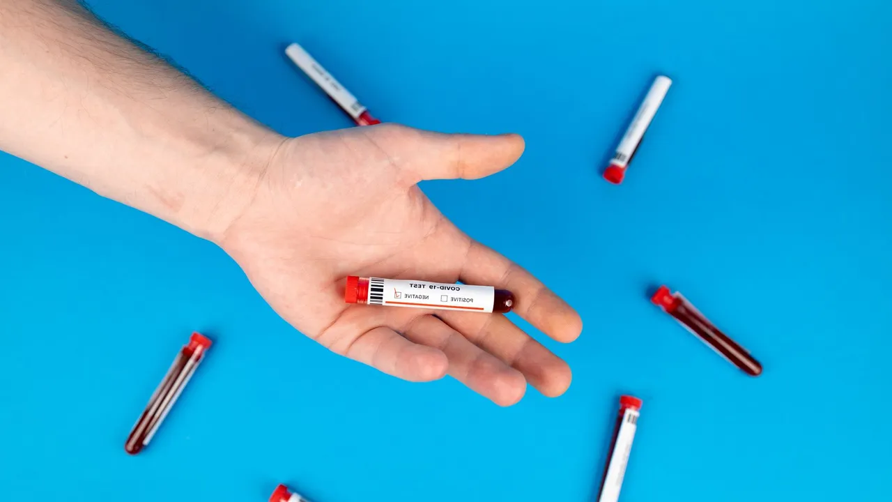 Close-up of a hand holding a blood sample vial on blue background, representing COVID-19 testing.