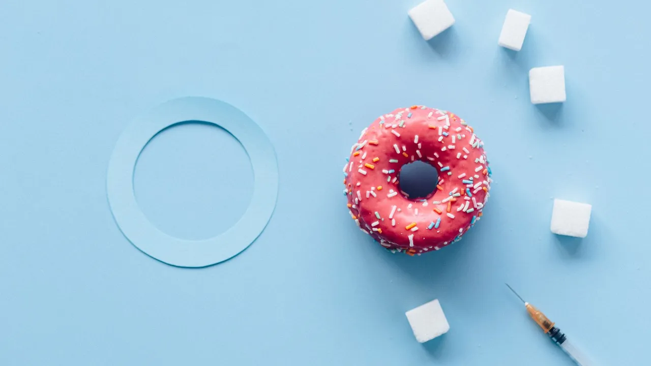 Flat lay of a pink donut, sugar cubes, and syringe on a blue background symbolizing diabetes awareness.