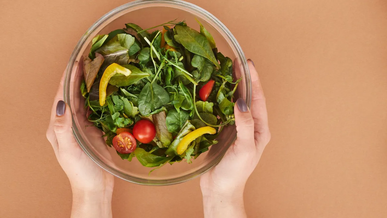Healthy fresh salad bowl with cherry tomatoes and greens held by hands on a brown surface.