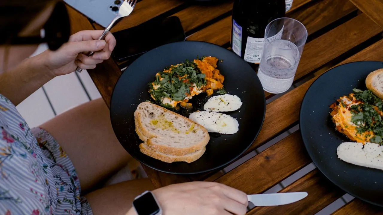 A casual outdoor brunch setting with fresh bread, cheese, and vegetables on dark plates.