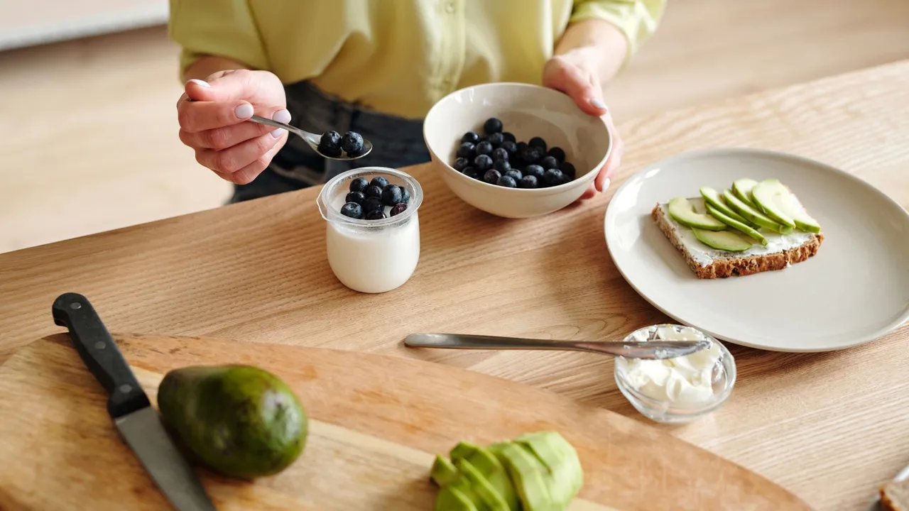 A person prepares a healthy breakfast with yogurt, blueberries, and avocado toast.