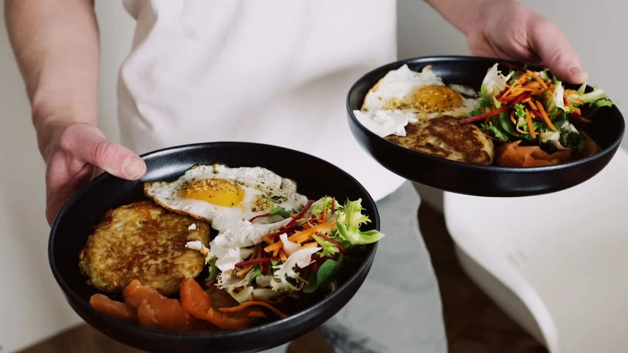 Faceless male in white clothes holding black plates with fried eggs and fresh salad for breakfast in cozy kitchen