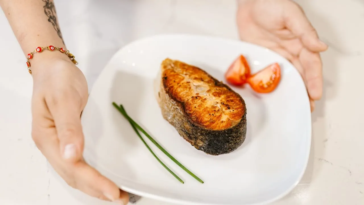 Close-up of grilled salmon served with tomatoes and chives on a plate, held by tattooed hands.