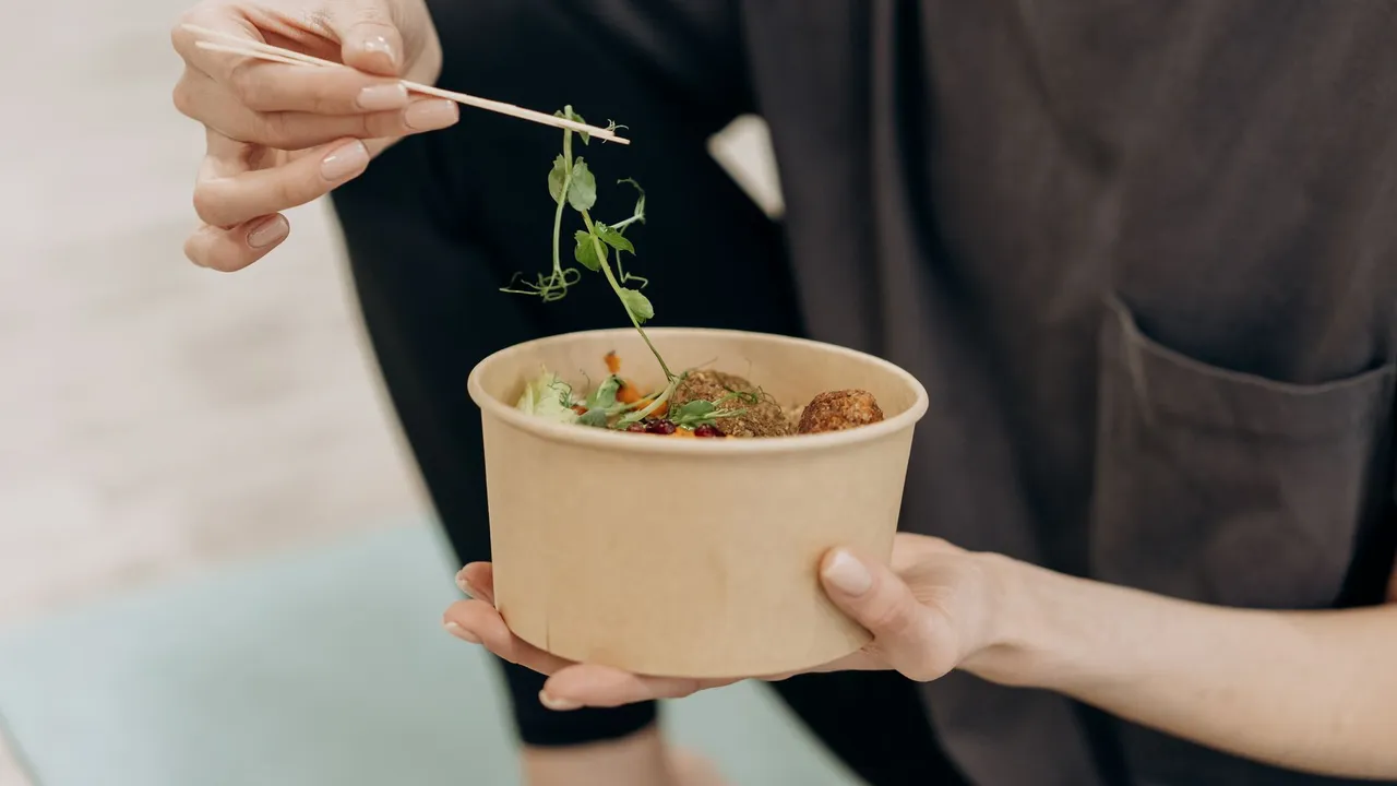 A woman holding a paper bowl filled with a healthy vegetarian meal in an indoor setting, promoting wellness.