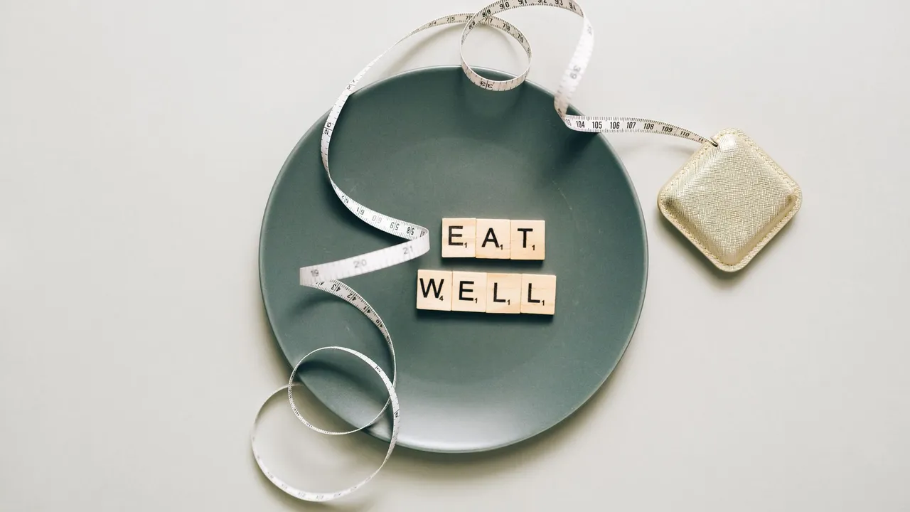Flat lay of scrabble tiles spelling 'eat well' on a plate with a tape measure, promoting healthy eating.