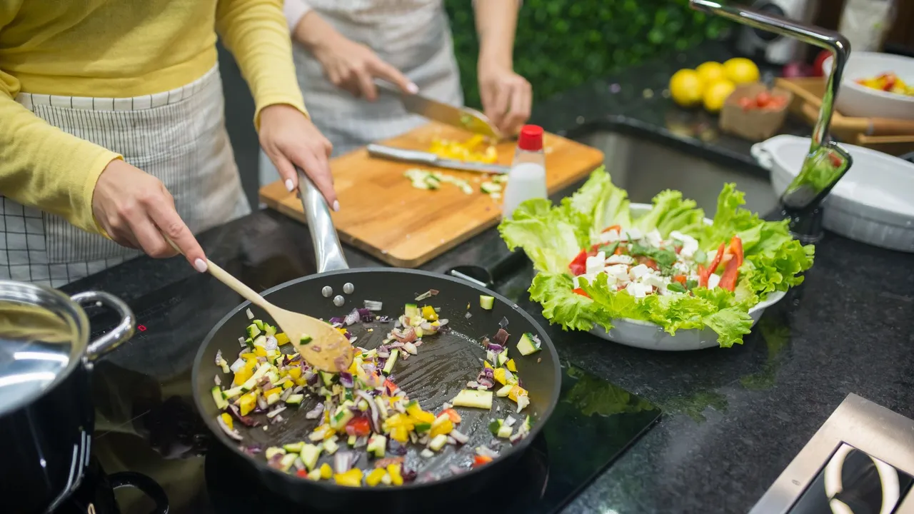Two adults cooking fresh vegetables on a stove, preparing a healthy meal in a modern kitchen.