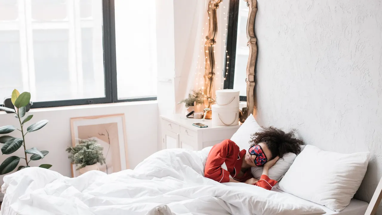 Woman wearing a sleep mask, resting on a cozy white bed surrounded by indoor plants in a bright bedroom.