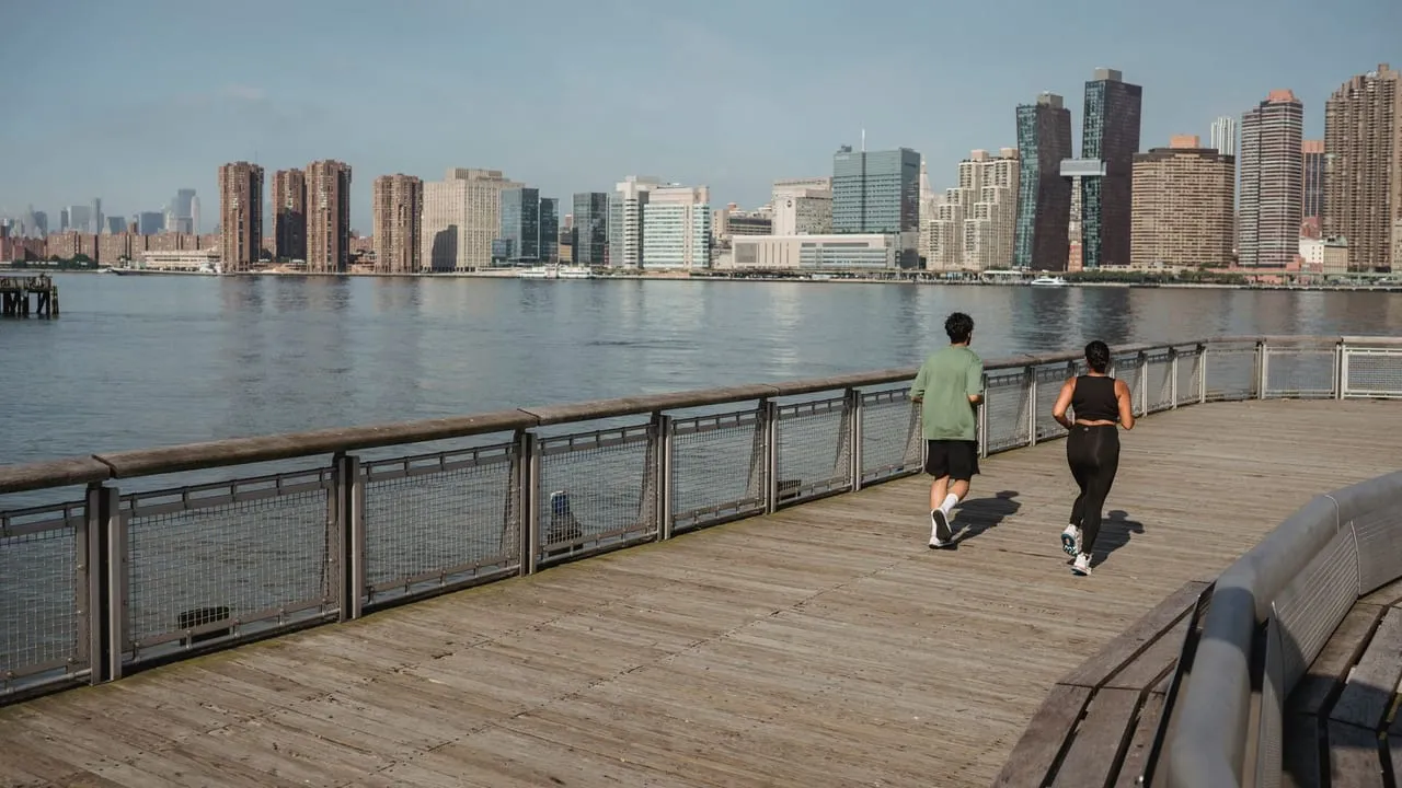 Two people jogging on a riverside path with a city skyline in the background.