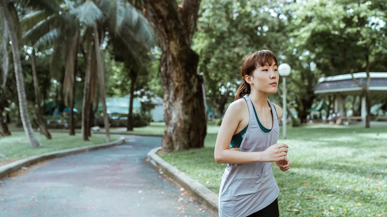 Thoughtful young Asian female athlete in sportswear running on asphalt road covered with faded leaves in city park in daytime and looking away