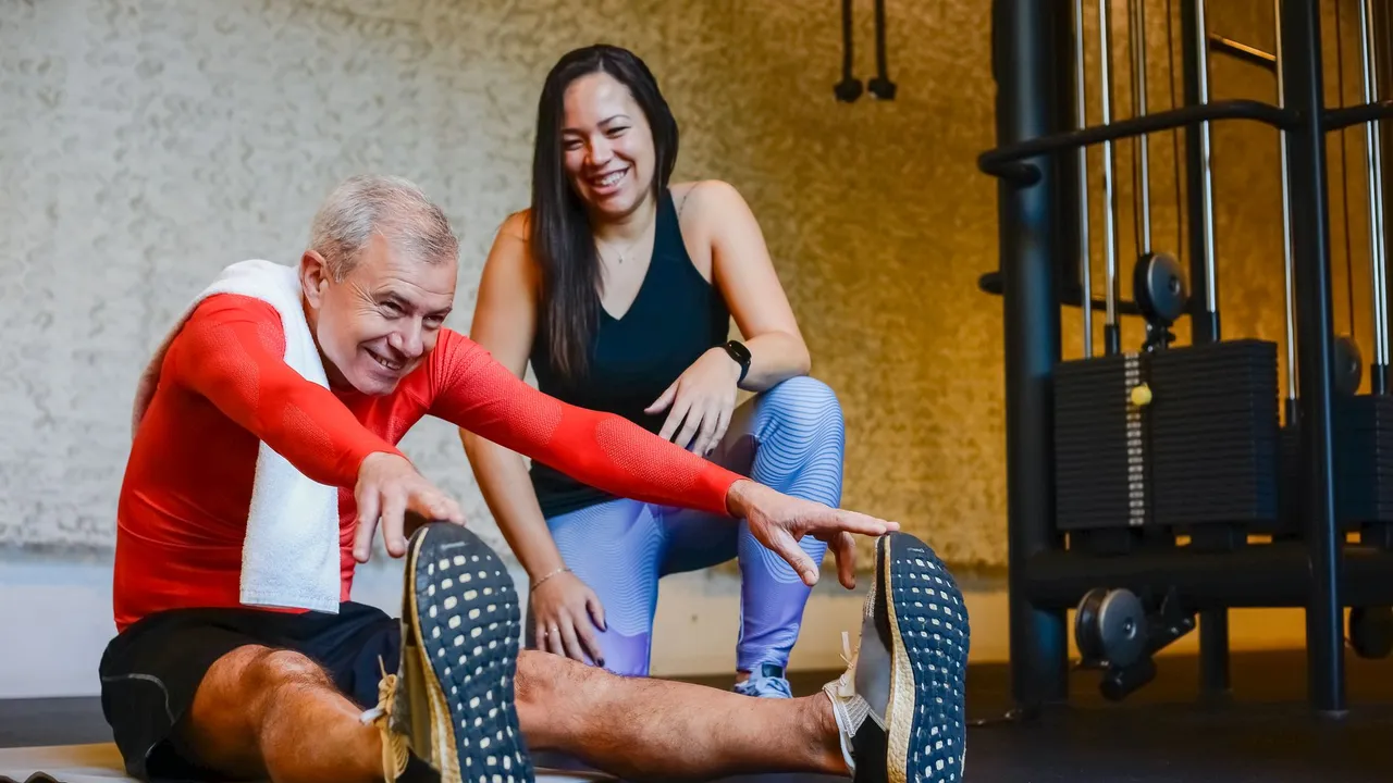 Elderly man in a gym stretching with a fitness coach, promoting a healthy lifestyle.