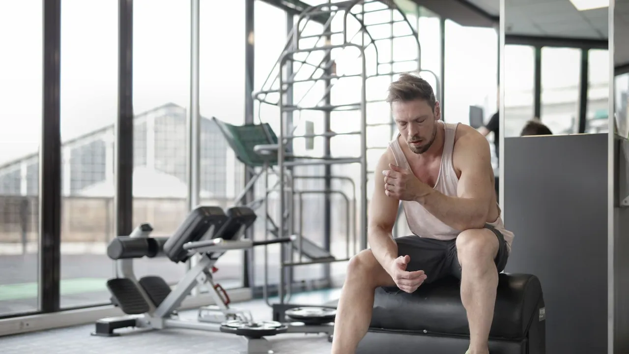 Tired young muscular male athlete in casual sportswear and sneakers sitting on bench and resting after intense weightlifting workout in modern fitness club