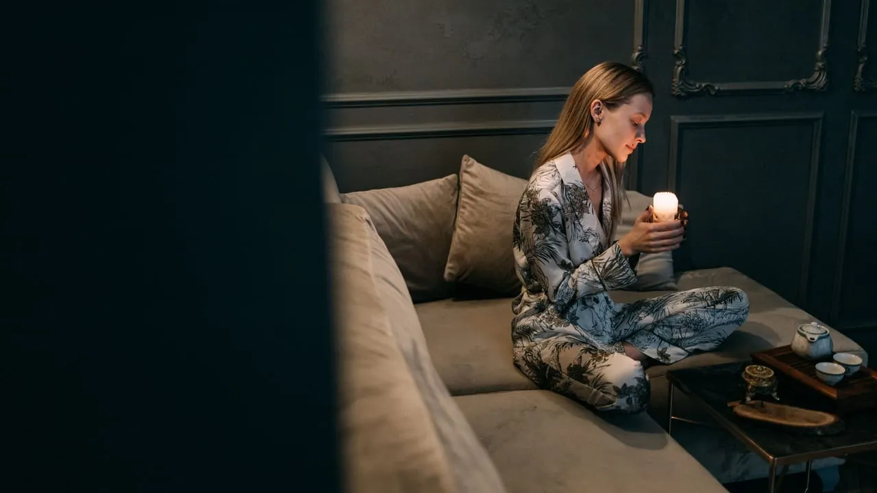 Woman in pajamas holding a candle, sitting on a cozy couch in a serene room.