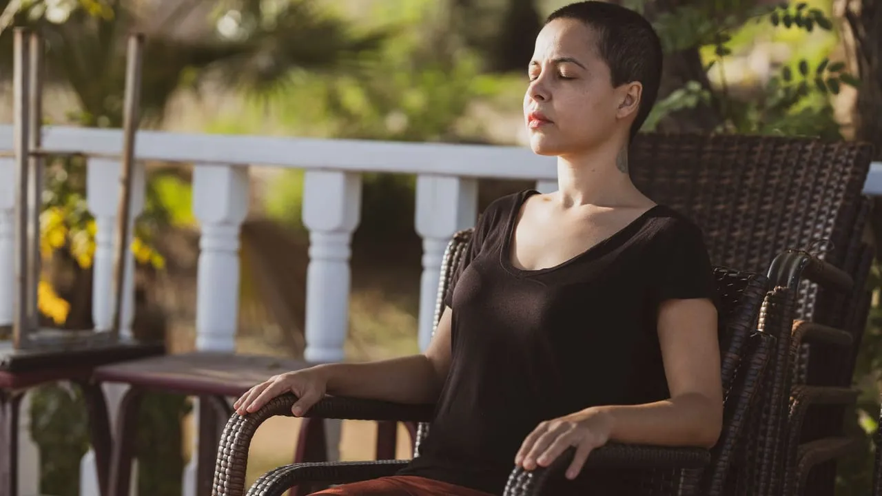 A woman in meditation with eyes closed sitting on a chair outdoors in the sunlight.