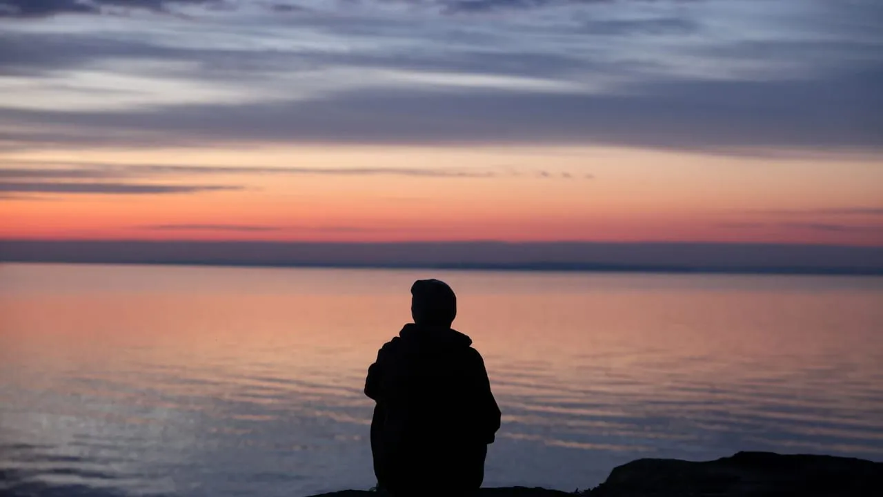 A contemplative silhouette of a person sitting by the calm sea during a beautiful sunset.