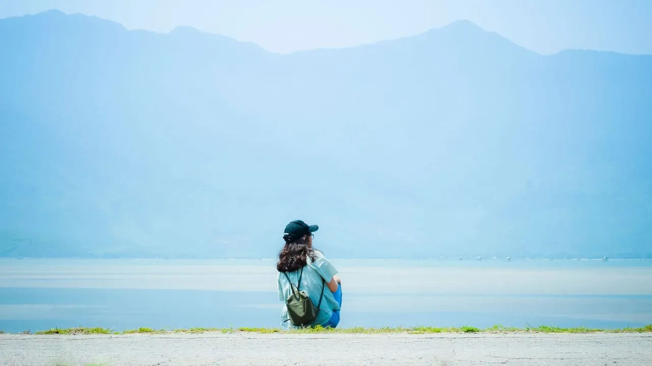 A person sitting alone by a lake, with mountains in the background, under a bright sky.