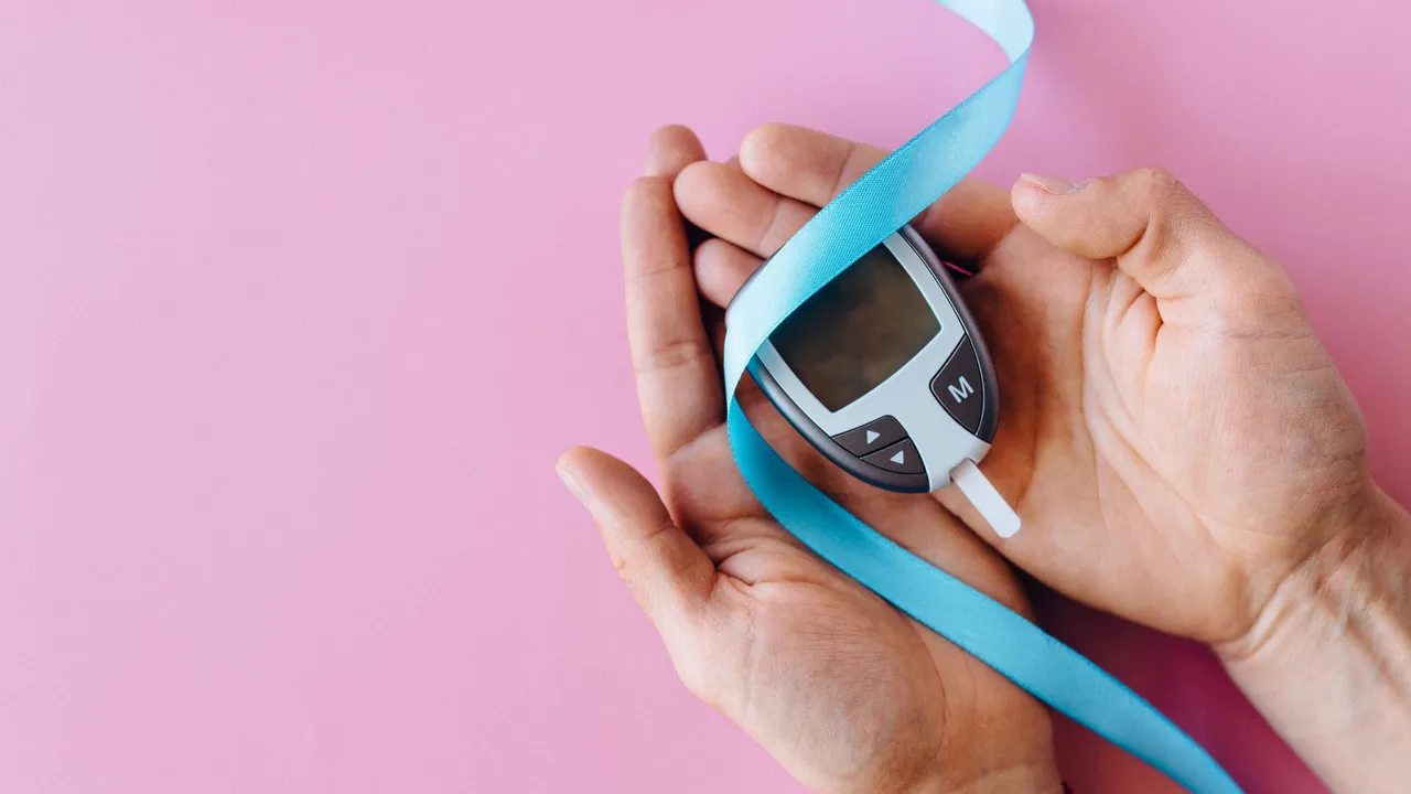 Hands holding a glucometer with a blue ribbon symbolizing diabetes awareness on a pink background.