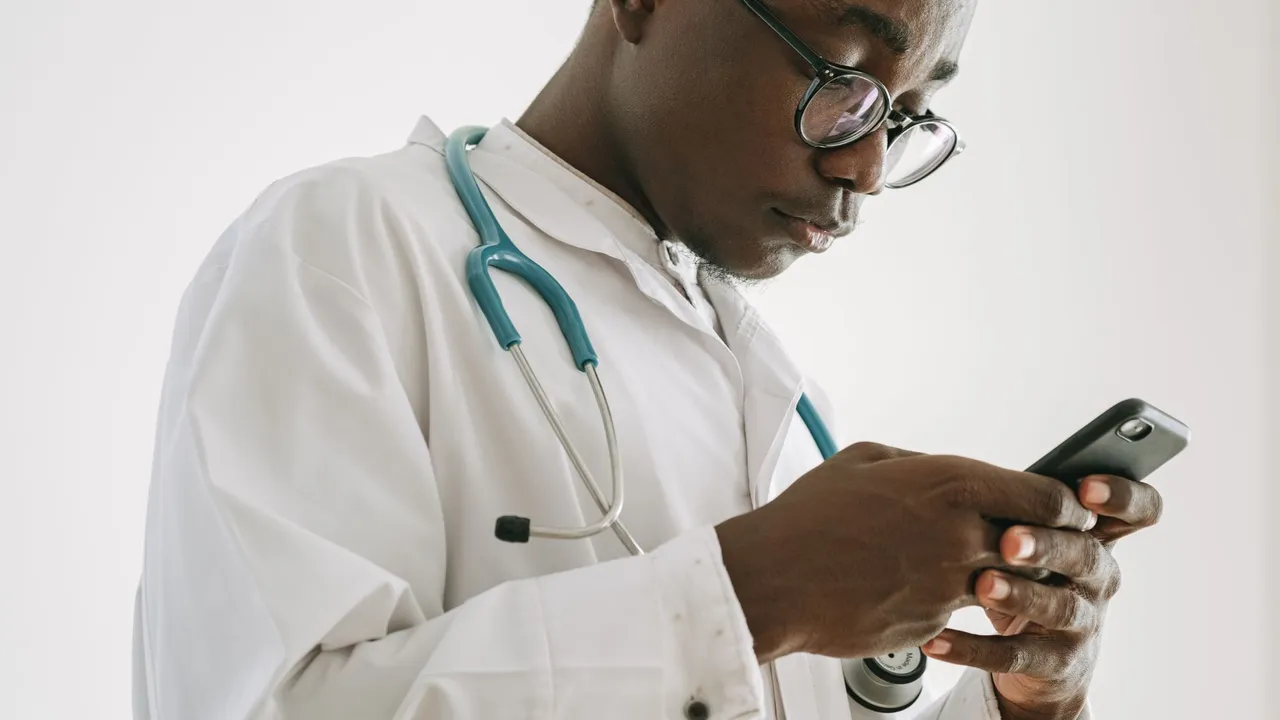 Focused doctor using smartphone, wearing a white coat and stethoscope, indoors.