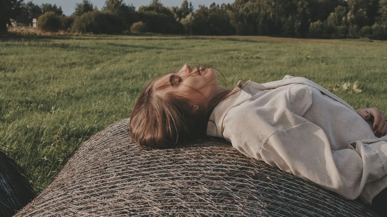 Woman lying on a hay bale in a sunny field, eyes closed, enjoying nature.