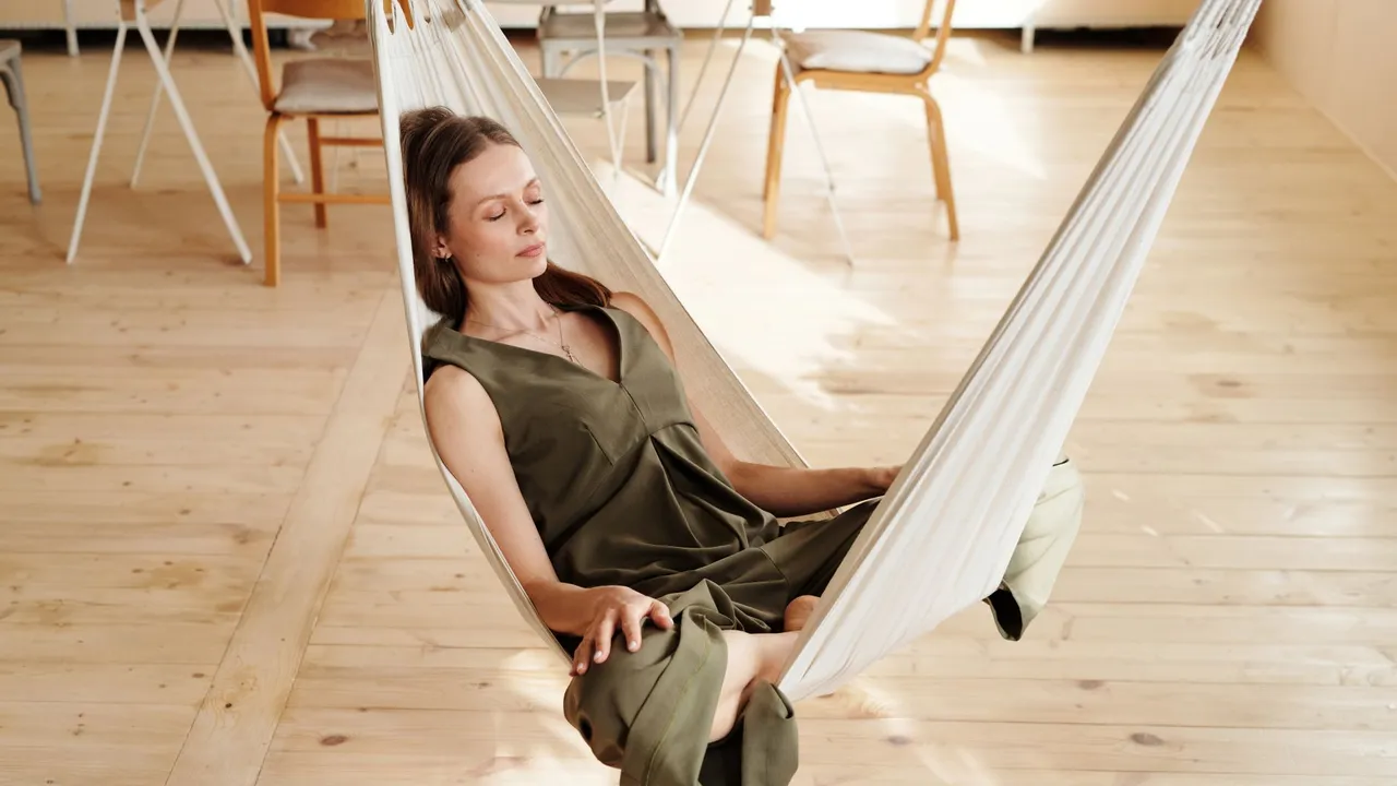 A woman in a green dress relaxes on a hammock inside a sunlit room with wooden floors.