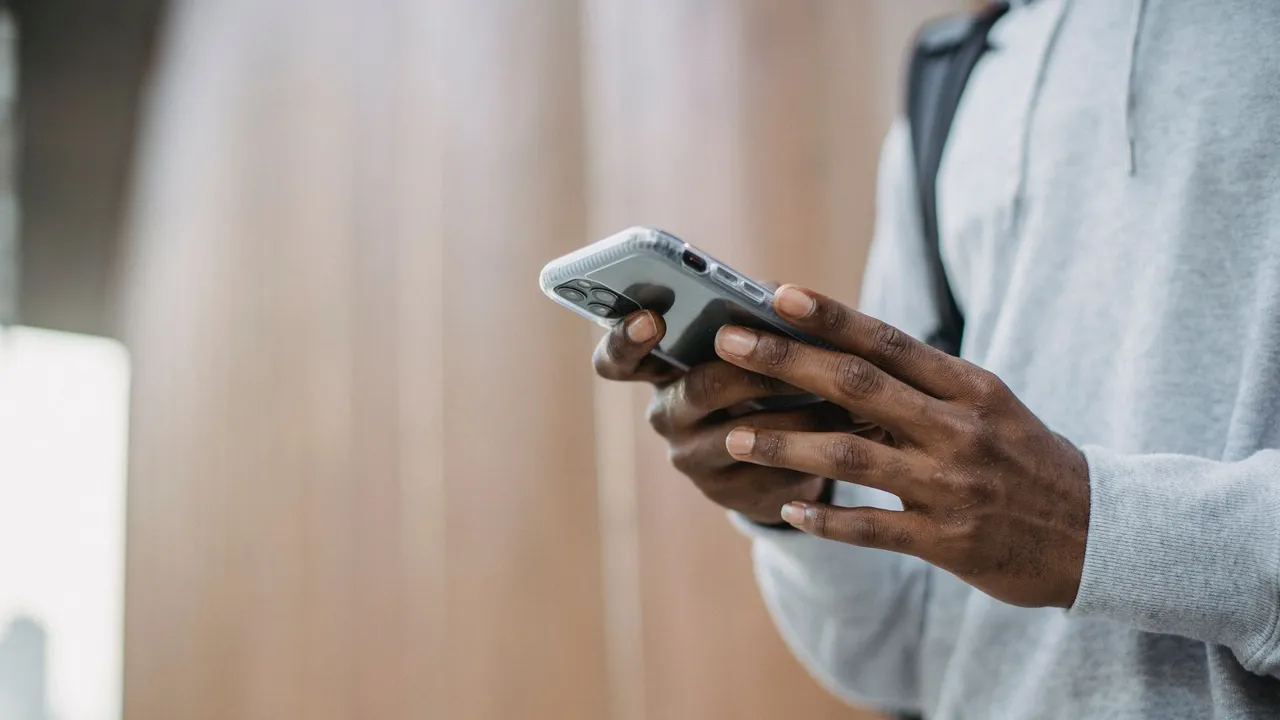 From below crop of unrecognizable African American young guy in casual clothes messaging on mobile phone while standing near wooden wall